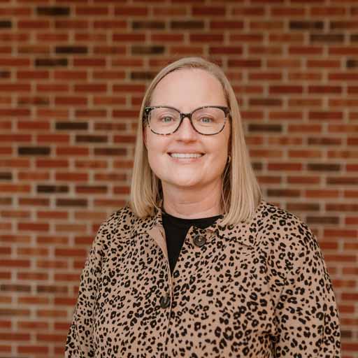 Woman with glasses and leopard print jacket smiles in front of brick wall